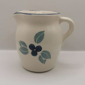 May include: Cream-colored ceramic pitcher with a blue rim and handle. The pitcher is decorated with a cluster of three dark blue berries and three light blue leaves. The pitcher is a classic design.
