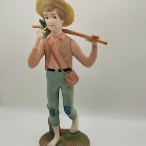 Può includere: Una statuetta in porcellana di un ragazzo con un cappello di paglia, una camicia rosa, un gilet marrone, pantaloni blu e un bastone sulla spalla. È scalzo e in piedi su una base verde.
