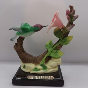 May include: A detailed figurine of a hummingbird in flight, approaching a pink flower. The bird has green and purple plumage, perched on a brown branch with green leaves. The base is a dark wood with a small metal label.