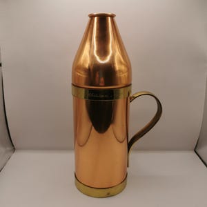 May include: A polished copper cocktail shaker with a conical top, a cylindrical body, and a brass handle. A brass band encircles the middle, featuring the engraved text "Christmas 1979". The base is finished with a matching brass rim.