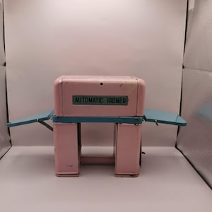 Puede incluir: Una planchadora automática de metal rosa y verde azulado vintage. La planchadora tiene una parte superior rectangular con las palabras "AUTOMATIC IRONER" en un rectángulo verde. Tiene dos mesas laterales de color verde azulado y está soportada por cuatro patas rosas.