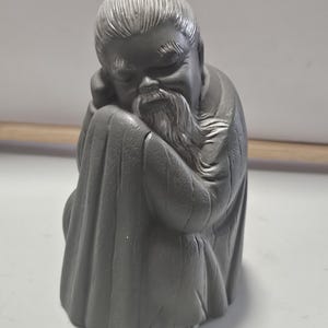 May include: A matte dark gray figurine of an elderly person with a long beard and hair tied in a topknot. The figure is depicted in a seated, hunched posture with eyes closed, wearing a robe with visible vertical folds and textured fabric details.