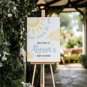 以下が含まれることがあります： 木製のイーゼルに立てられた白い看板。テキストは「Here comes the son」と「Welcome to Alyssa's Baby Shower 06.08.2025」です。黄色い太陽と雲のイラスト、青い文字が特徴です。
