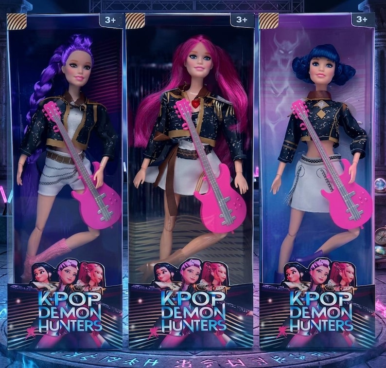May include: Three KPOP Demon Hunters dolls in clear packaging. Each doll has a different hair colour: purple, pink, and blue. They wear black jackets, white skirts, and hold pink electric guitars. The packaging features the text "KPOP DEMON HUNTERS".