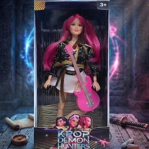 May include: A doll with pink hair and a pink guitar, packaged in a box. The doll wears a black jacket with gold accents, a white skirt, and a brown belt. The box features the text "KPOP DEMON HUNTERS" and the age range "3+".