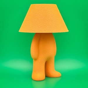 Op de afbeelding: Oranje tafellamp met een textuuroppervlak, gevormd als een persoon. De lamp heeft een conische lampenkap en een lichaam met benen. De achtergrond is effen groen.