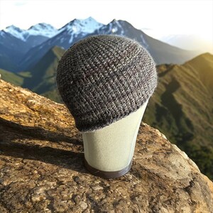 Puede incluir: Un gorro de punto gris con sutiles motas rojas y blancas, exhibido en una cabeza de maniquí. El gorro se encuentra sobre un fondo de una cordillera y una superficie rocosa.