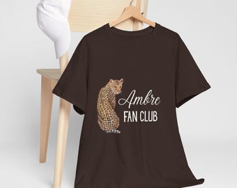 May include: A brown t-shirt featuring a leopard illustration and the white text "Ambre FAN CLUB". The t-shirt is displayed on a wooden hanger. A white baseball cap is placed on a wooden chair.