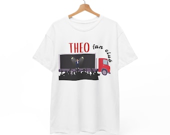 May include: White t-shirt featuring a stage scene on a red truck, with the text "THEO fan club" in red. The stage shows a person under spotlights and a crowd. The t-shirt is displayed on a wooden hanger.