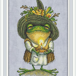 May include: Cross-stitch image of a green frog wearing a witch's hat adorned with flowers and leaves. The frog is holding a small cake with a lit candle. The frog is wearing a white apron and a brown dress.