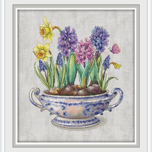 May include: Cross-stitch artwork of a floral arrangement in a decorative blue and white planter. The arrangement features yellow daffodils, purple and pink hyacinths, and blue grape hyacinths. A small pink butterfly is also included in the design.