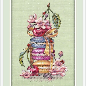 May include: A cross-stitch embroidery pattern featuring a vertical stack of three colorful macarons in blue, pink, and yellow, tied with a gold ribbon. The design includes red cherries, green leaves, pink cherry blossoms, and small ladybugs.