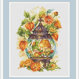Könnte beinhalten: Kreuzstich-Kunstwerk mit einer dekorativen Laterne mit Glaskugel, in der sich zwei Zwerge und ein Bienenstock befinden. Die Laterne ist von orangefarbenen Blumen, grünen Blättern und orangefarbenen Lampionfrüchten umgeben, vor hellem Hintergrund.