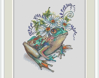Può includere: Ricamo a punto croce di una rana colorata con una corona di fiori di margherita. La rana ha un corpo verde e blu con accenti arancioni. L'opera è incorniciata in una cornice bianca.
