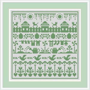 May include: A framed cross-stitch sampler featuring a green and white folk art design. The design includes houses, trees, birds, and floral elements. The artwork is framed in a light green mat and white frame.