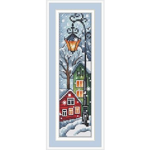 以下が含まれることがあります： 雪に覆われた赤い家と緑の家、背の高い黒い街灯が描かれた縦型のクロスステッチ刺繍パターン。このシーンには、オレンジ色に輝くランタン、葉のない木々、青い空に舞う白い雪の結晶が表現されています。