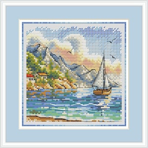 May include: Cross-stitch artwork depicting a coastal scene with a sailboat on the water. The artwork features a blue sky, mountains, and a small house on the shore. The piece is framed in a light blue border.
