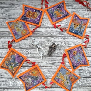 May include: A Halloween-themed garland featuring eight pennant-shaped cross-stitch panels with orange borders, depicting pumpkins, ghosts, a haunted house, a witch hat, a black cat, and eyeballs, accompanied by a small plastic skeleton and coffin.