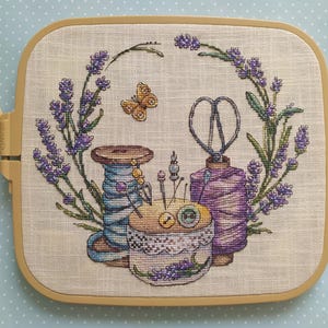 Peut inclure: Broderie au point de croix représentant une couronne de lavande entourant des outils de couture. Un papillon, des ciseaux, des bobines de fil et un coussin à épingles sont représentés. La broderie est dans un cercle carré, beige, avec un fond à pois bleu clair.