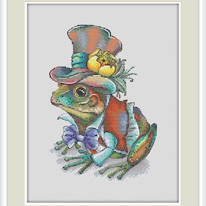 May include: A cross-stitch design of a frog wearing a colorful top hat, bow tie, and vest. The frog is green, orange, and blue, with a yellow flower on the hat. The image is framed in a white border.