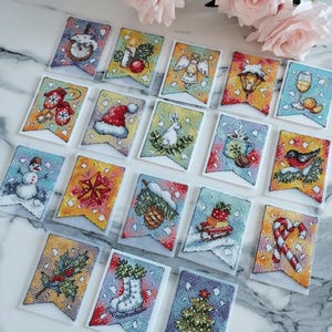 Peut inclure: Une collection de bannières de Noël brodées au point de croix. Chaque bannière présente un motif de vacances unique, comme des bonhommes de neige, des anges et des décorations festives. Les bannières sont disposées sur une surface blanche avec des roses roses en arrière-plan.