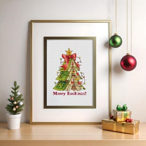 May include: Cross-stitch art of a Christmas tree made of books, adorned with a red bow and star. The phrase "Merry Bookmas!" is stitched below. The framed artwork is displayed with a small tree and ornaments.