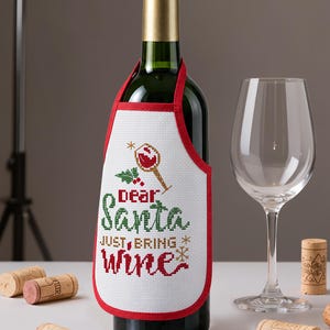 May include: A dark green wine bottle wearing a white fabric apron with red trim. The apron features cross-stitch embroidery of a wine glass, holly leaves, and the text "Dear Santa JUST, BRING wine" in red and green lettering with small gold stars.