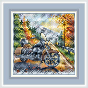 May include: A cross-stitch embroidery depicting a dark motorcycle parked on a road during autumn. The scene features vibrant yellow and orange trees, a mountain backdrop, and a guardrail. The artwork is enclosed in a light blue frame.