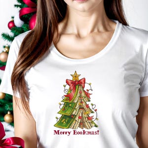 May include: White t-shirt featuring a Christmas tree design made of books. The tree is decorated with a red bow, a gold star, and string lights. The text "Merry Bookmas!" is printed below the tree.