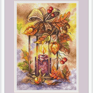 Könnte beinhalten: Kreuzstich-Kunstwerk einer Laterne mit einer brennenden lila Kerze. Die Laterne ist mit Herbstblättern, Beeren und einer Schleife verziert. Das Kunstwerk ist in einem weißen und lavendelfarbenen Rahmen gerahmt.
