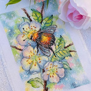 Può includere: Ricamo a punto croce raffigurante una farfalla con ali arancioni, nere e gialle, posata su un ramo fiorito. I fiori sono bianchi e azzurri, con foglie verdi. Lo sfondo è una morbida miscela di colori.