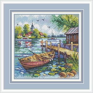 May include: A cross-stitch embroidery depicting a lakeside scene with a wooden rowboat moored at a dock. The landscape features a small cabin, a distant village with a church spire, green trees, and birds in the sky, all framed in a light blue border.