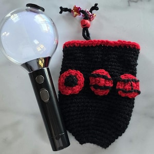 May include: A black and red crocheted light stick cover with three red and black decorative accents. The cover is next to a black and silver light stick with a clear, spherical top. A decorative string with star-shaped beads is attached to the cover.