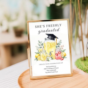 May include: A rectangular gold-framed sign featuring the text SHE'S FRESHLY graduated and CREATE YOUR CELEBRATION SIP with instructions. The graphic shows a large lemonade dispenser with a graduation cap, lemons, and strawberry-infused drinks.