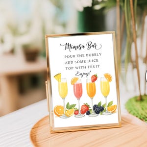Könnte beinhalten: Ein goldgerahmtes Schild mit der Aufschrift "Mimosa Bar" in Schreibschrift. Der Text lautet: "Gießen Sie den Sekt ein, fügen Sie Saft hinzu, belegen Sie ihn mit Früchten, genießen Sie es!" Aquarellillustrationen von Mimosen und Früchten.