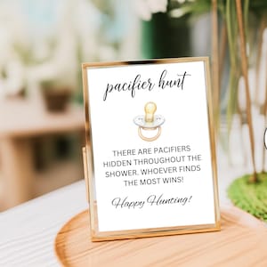May include: A gold-framed sign with the words "pacifier hunt" at the top. The sign includes a drawing of a pacifier and text that reads "There are pacifiers hidden throughout the shower. Whoever finds the most wins! Happy Hunting!"