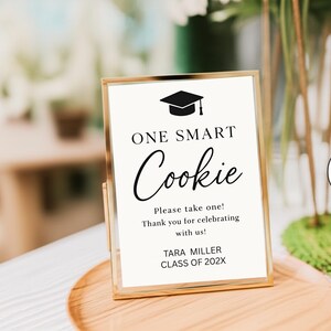 May include: A vertical rectangular sign in a gold metal frame featuring a black graduation cap icon and the text: ONE SMART Cookie, Please take one! Thank you for celebrating with us! TARA MILLER CLASS OF 202X, printed in black on a white background.