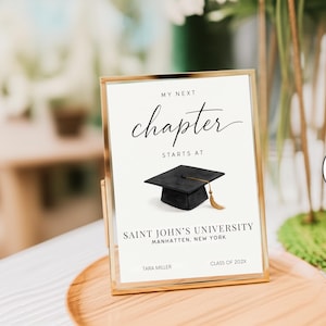 May include: A rectangular gold-toned frame holds a white sign with black text that reads MY NEXT chapter STARTS AT, featuring an illustration of a black graduation cap with a gold tassel, followed by SAINT JOHN'S UNIVERSITY, MANHATTEN, NEW YORK, TARA MILLER, CLASS OF 202X.