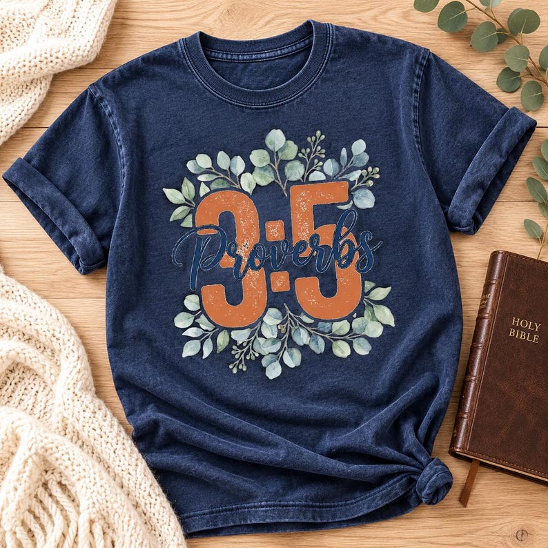 May include: A navy blue short-sleeve t-shirt featuring a graphic print of the text "3:5" in large orange numbers and the word "Proverbs" in dark blue script, surrounded by a circular arrangement of green eucalyptus leaves and branches.