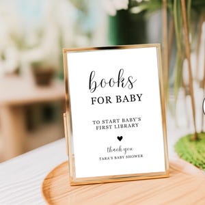May include: A gold-framed sign with the words "books FOR BABY" in elegant black script. Below, it reads "TO START BABY'S FIRST LIBRARY" and "thank you TARA'S BABY SHOWER". The sign is on a wooden tray.
