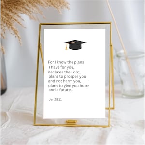 May include: A gold-framed print featuring a graduation cap illustration and the text "For I know the plans I have for you..." from Jeremiah 29:11. The print is displayed on a white surface, with a blurred background.