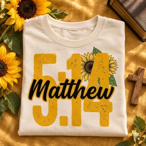 May include: A cream-colored t-shirt featuring a large yellow graphic with the text 5:14 and Matthew in black script. The design includes a detailed illustration of a sunflower with green leaves integrated into the numbers and text.