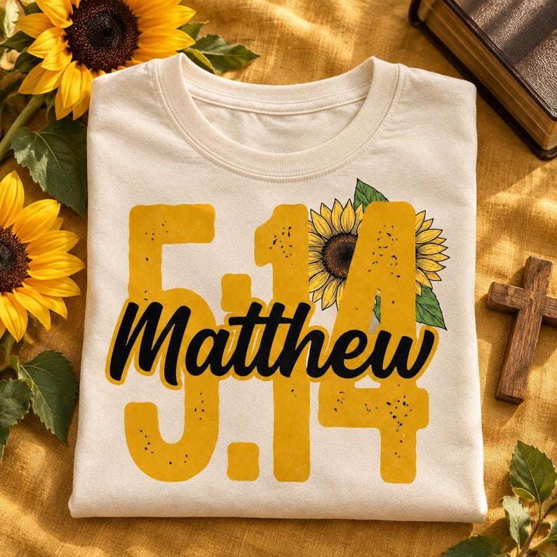 May include: A cream-colored t-shirt featuring a large yellow graphic with the text 5:14 and Matthew in black script. The design includes a detailed illustration of a sunflower with green leaves integrated into the numbers and text.