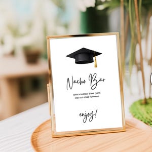 May include: A vertical rectangular gold-toned metal frame containing a white sign with a black graduation cap graphic. The text reads: Nacho Bar, GRAB YOURSELF SOME CHIPS AND ADD SOME TOPPINGS, and enjoy! The sign is displayed on a wooden surface.
