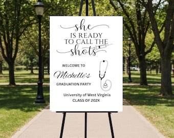 May include: A white graduation party sign on a black easel. The sign reads "She is ready to call the shots" with a syringe graphic. Below, it says "Welcome to Michelle's Graduation Party" and "University of West Virginia, Class of 202X". A stethoscope graphic is also included.