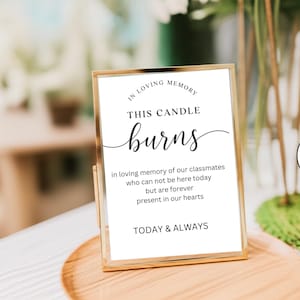 Op de afbeelding: Een goudkleurig omlijst bord met de tekst "IN LOVING MEMORY THIS CANDLE burns" in elegant schrift. Het bord bevat een herinneringsboodschap: "in loving memory of our classmates who can not be here today but are forever present in our hearts. TODAY & ALWAYS."