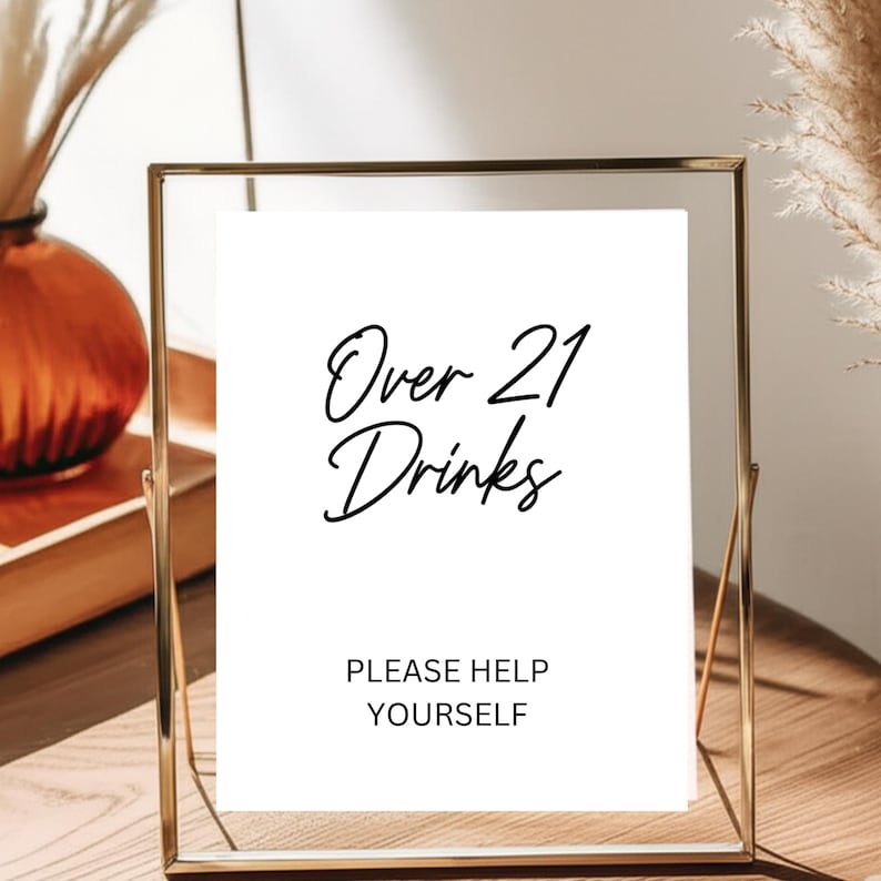 Op de afbeelding: Een goudkleurig ingelijst bord met de tekst "Over 21 Drinks" in zwart script. Daaronder staat "PLEASE HELP YOURSELF". De achtergrond is een neutraal beige, ideaal voor een feest of evenement.