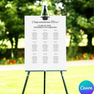 May include: A white graduation seating chart on a black easel. The chart reads "Congratulations Tara!" and lists names under table numbers. A graduation cap graphic is at the top. The background is a blurred green outdoor scene.