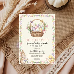 May include: Easter-themed invitation with a watercolor illustration. A woven basket filled with decorated eggs and flowers is at the top. The text "HOP ON OVER for an Easter brunch and egg hunt" is displayed. A bunny and chick are at the bottom.