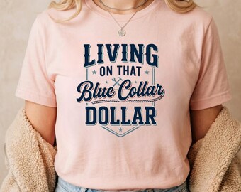 Puede incluir: Una camiseta de manga corta de color rosa claro con texto azul oscuro que dice "LIVING ON THAT Blue Collar DOLLAR", un pequeño icono de llave inglesa y un gráfico de cuerda, enmarcado por un borde azul fino con estrellas sobre tela rosa.